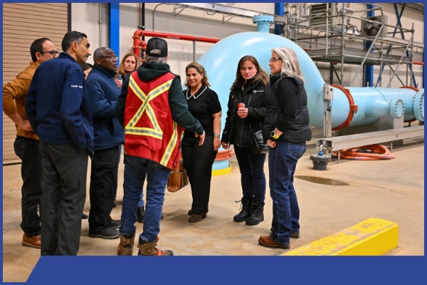 A group of people stand inside an industrial facility while a worker in a red safety vest explains equipment. Large blue machinery, pipes, and safety barriers surround the group in the well‑lit space.