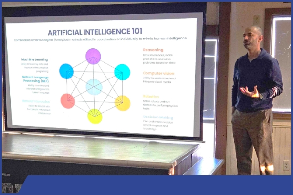 A male speaker stands besides a projection of a presentation centered on AI